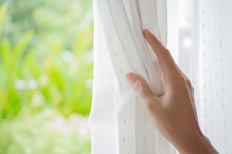 Woman's hand opening curtains in the bedroom with natural light and garden background, Spring Money-Saving Tips