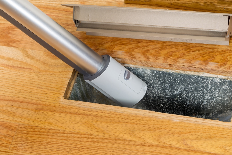Horizontal photo of vacuum cleaning inside heater floor vent with Red Oak Floors in background. 7 Furnace Maintenance Tips.